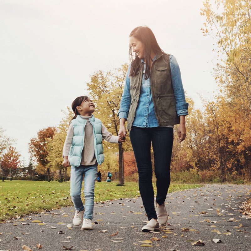 mother and daughter holding hands walking on path with fall color trees in background